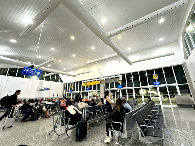 Sala de embarque do Aeroporto Glauber Rocha em Vitória da Conquista. A foto mostra um salão amplo com fileiras de cadeiras cinzas, teto alto com tubulação aparente e grandes janelas de vidro com vista para a pista à noite.