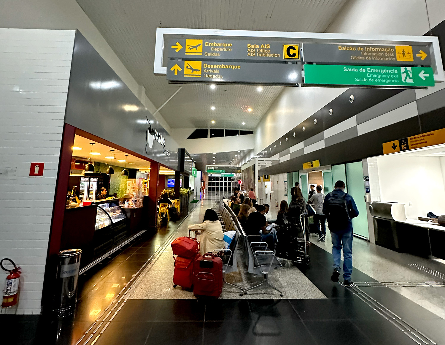 Saguão de entrada do Aeroporto Glauber Rocha em Vitória da Conquista. A imagem mostra o interior do terminal com passageiros aguardando, lanchonete à esquerda e placas de sinalização de embarque e desembarque.