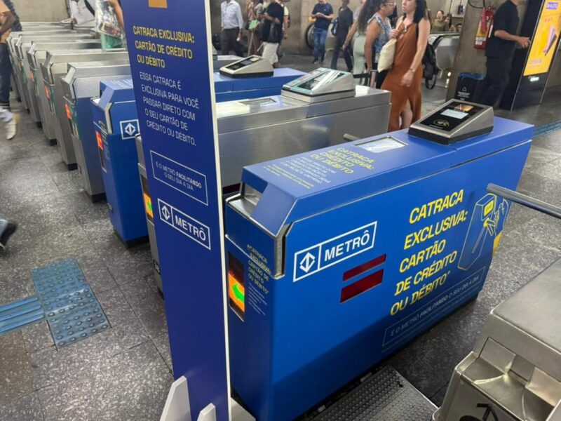 Catraca de metrô na cor azul com adesivos amarelos indicando "Catraca Exclusiva: Cartão de Crédito ou Débito" e o logotipo do Metrô de São Paulo. Ao lado, fileiras de catracas comuns prateadas e passageiros ao fundo.