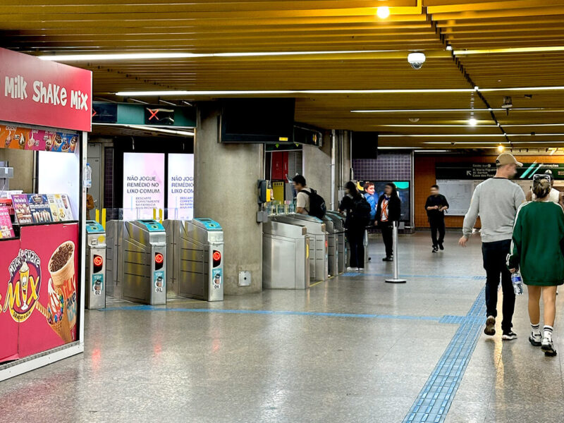 Passageiros caminhando em direção às catracas de acesso da estação Brigadeiro do Metrô SP. Ao lado, quiosque de alimentação vermelho.