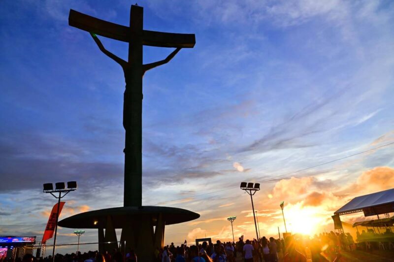 Silhueta da estátua do Cristo crucificado de Mário Cravo Jr. em Vitória da Conquista contra um céu de pôr do sol azul e alaranjado. A imagem mostra a movimentação de turistas na base da Serra do Periperi no fim da tarde.