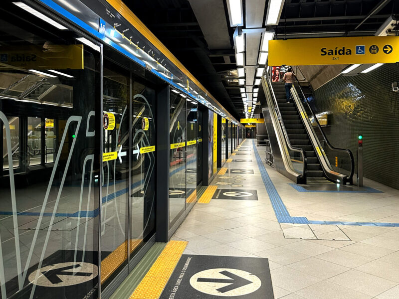 Plataforma de embarque de estação do Metrô de São Paulo moderna, com portas automáticas de vidro, piso de granito e sinalização amarela de saída.