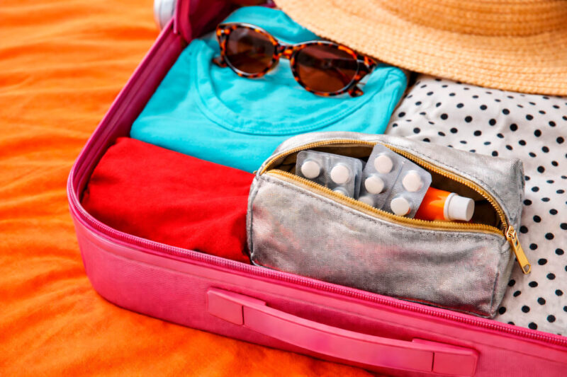 Mala de viagem rosa aberta com roupas coloridas dobradas. Sobre as roupas, há uma necessaire prateada pequena com cartelas de remédios à mostra e um tubo de pomada. Um chapéu de palha e óculos de sol estão ao fundo.