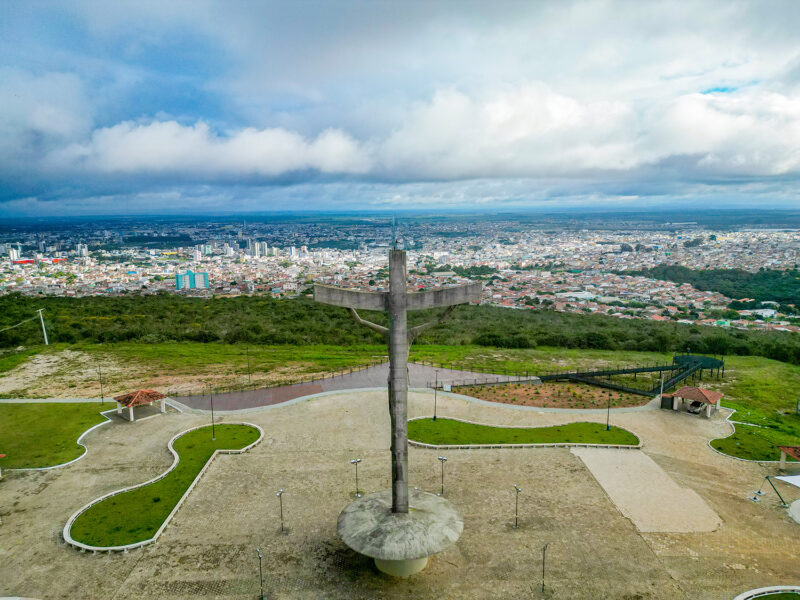 Vista aérea traseira da estátua do Cristo de Mário Cravo no alto da Serra do Periperi, com a cidade de Vitória da Conquista ao fundo sob um céu nublado.