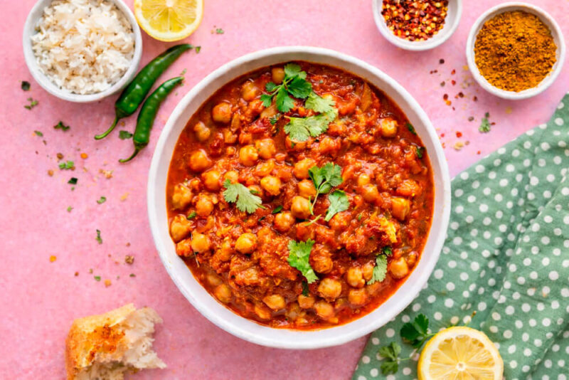 Tigela branca com Chana Masala (curry de grão-de-bico) em molho vermelho intenso, decorado com folhas de coentro fresco. Ao redor, potes pequenos com arroz branco e especiarias em pó, pimentas verdes inteiras e um pedaço de pão naan sobre um fundo rosa.