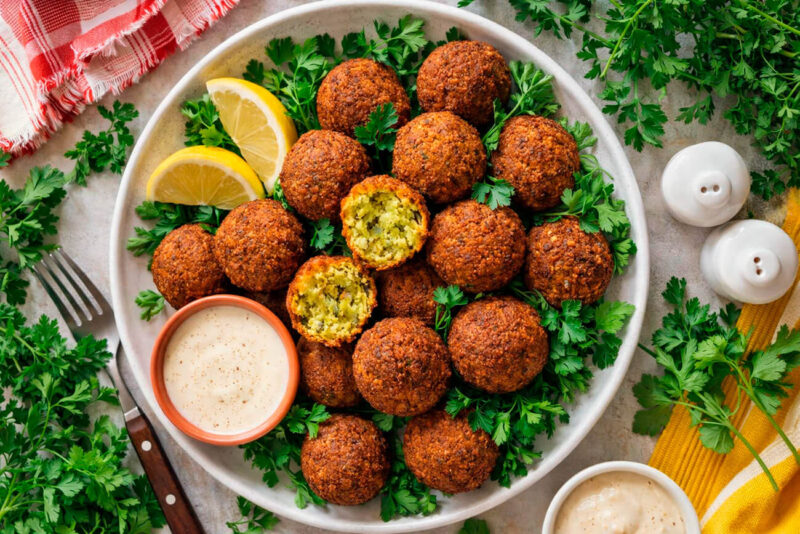 Prato de cerâmica branca servido com vários bolinhos de falafel fritos e dourados, decorados com ramos de salsa fresca e fatias de limão siciliano. Ao lado, dois potes pequenos com molhos cremosos (tahine e homus).