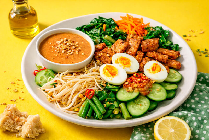 Prato branco grande contendo Gado-Gado (salada indonésia) com ovos cozidos, tempeh frito, feijão-vagem, pepino e brotos. Ao centro, um pote com molho de amendoim cremoso. O fundo da foto é amarelo vibrante.