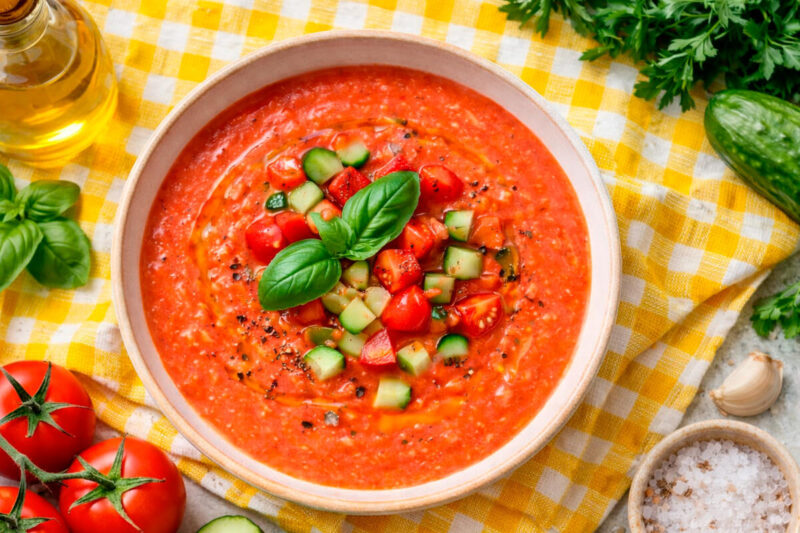 Tigela de sopa fria de tomate (Gaspacho) em cor vermelho vibrante, decorada com cubinhos de pepino, tomate e folhas de manjericão fresco. Ao fundo, ingredientes crus como tomates e azeite sobre uma toalha xadrez amarela.