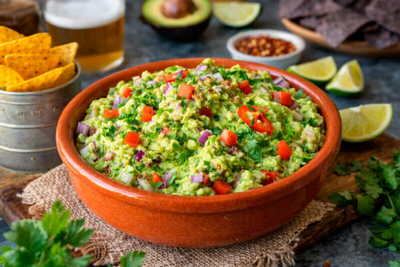 Tigela de barro rústica cheia de guacamole fresco, decorado com cubos de cebola roxa, fatias de pimenta vermelha, coentro e especiarias. Ao lado, um copo de metal com chips de tortilla (nachos) e fatias de limão sobre uma mesa escura.