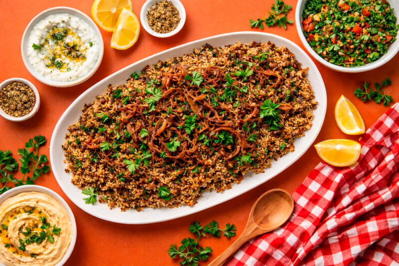 Travessa oval grande servida com Mujadara (mistura de arroz e lentilhas escurecidas), coberta com uma camada generosa de cebolas fritas crocantes e salsinha fresca. Ao redor, potes com acompanhamentos como homus, coalhada seca e salada picada sobre um fundo laranja vibrante.