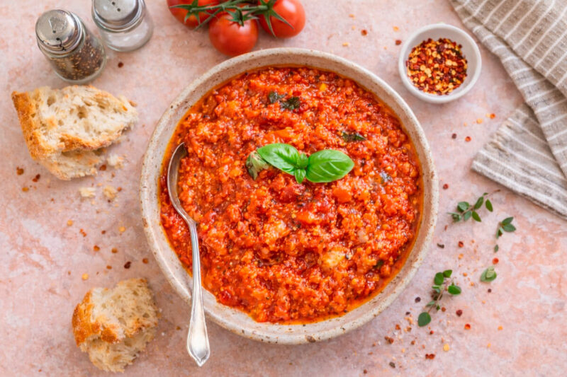 Tigela rústica com Pappa al Pomodoro (sopa toscana densa de pão e tomate), decorada com folhas frescas de manjericão. Ao redor, pedaços de pão italiano rústico, um potinho com pimenta calabresa e tomates frescos no ramo sobre uma superfície de pedra rosada.