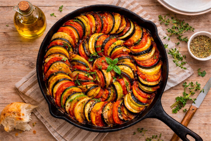 Panela de ferro contendo Ratatouille, com fatias finas de abobrinha amarela e verde, berinjela e tomate organizadas em espiral perfeita. A mesa de madeira rústica ao redor contém azeite, pão e ramos de tomilho fresco.