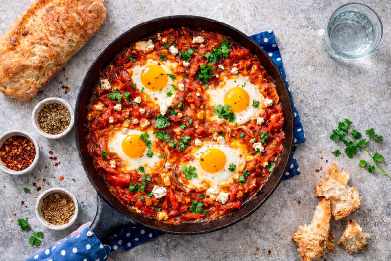 Frigideira de ferro preta contendo Shakshuka: quatro ovos com gemas moles cozidos em molho de tomate vermelho e pedaçudo, salpicados com queijo feta esfarelado e salsinha verde. Ao lado, um pão rústico cortado, potinhos de especiarias e um guardanapo azul com bolinhas.